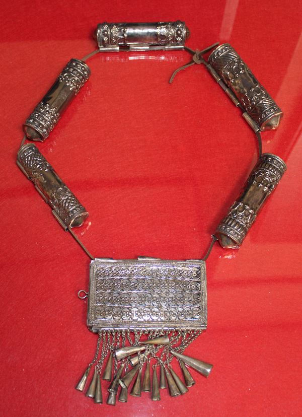 photo of an elaborate silver necklace consisting of long cylindrical beads and a rectangular pendant with a row of silver cone-shaped bells suspended from it by fine chains. Every part is decorated with intricate embossed and filigreed designs.